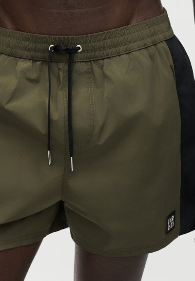 Homme portant un short de bain vert olive et noir avec un cordon noir et un petit patch logo sur la jambe gauche.