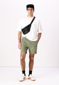Weißes T-Shirt mit Blumenmuster, olivgrüne Shorts, schwarze Gürteltasche, schwarze Socken und weiße Schuhe mit roten Streifen. Schwarze Kappe.
