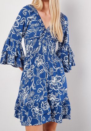 Kobieta w niebieskiej sukience w kwiaty z białymi wzorami, z marszczonym stanem, dekoltem w serek oraz falbanianymi rękawami i dołem w kilku warstwach.