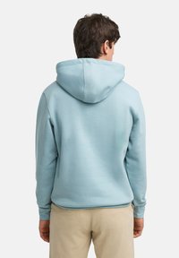 Felpa in cotone azzurro chiaro con cappuccio regolabile, polsini e orlo a costine, vista di spalle, indossata con pantaloni beige.