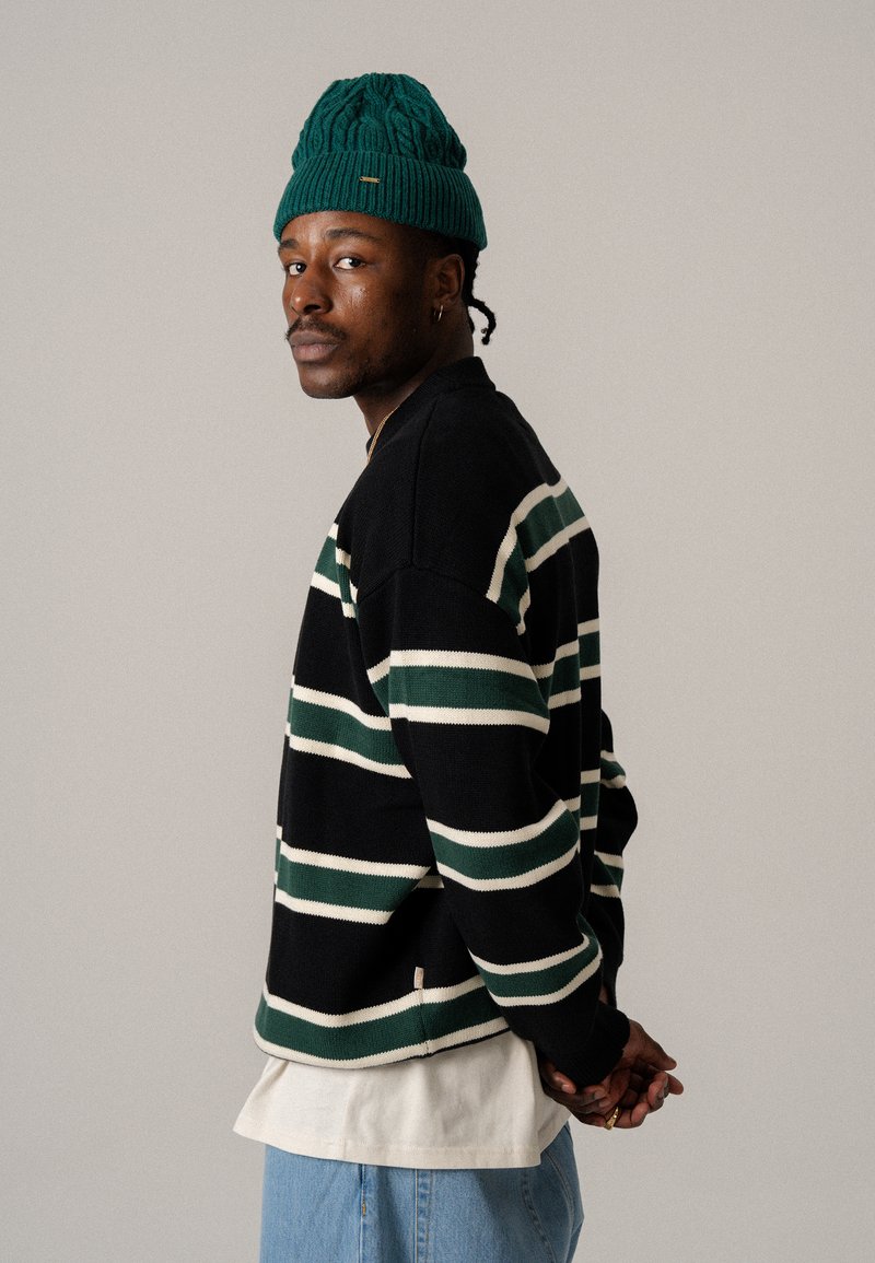 Zwart en groen gestreepte sweater met witte accenten, ronde hals, gedragen over een witte shirt, gecombineerd met lichtblauwe denim. Gebreide groene beanie.