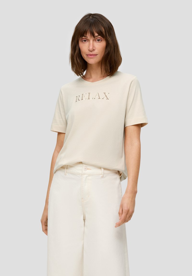 Hellbeige Kurzarm-T-Shirt mit dezent gesticktem "RELAX"-Schriftzug. Weiches Baumwollmaterial, kombiniert mit weißen weiten Hosen.