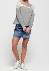 Grauer Pullover mit Spitzenakzent an den Schultern, kombiniert mit einem blauen Jeansrock. Das Model trägt weiße Sneakers und hat die Arme verschränkt.
