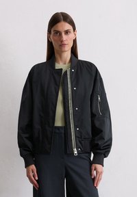Black bomber jacket with a smooth synthetic fabric. Features ribbed cuffs, a zip pocket on the sleeve, and silver snaps and zipper.