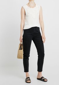 Top bianco senza maniche con dettagli in pizzo, abbinato a jeans skinny neri. La modella tiene in mano una borsa intrecciata e indossa sandali neri con borchie.