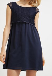 Robe bleu marine avec un haut en tricot présentant un design à bords ondulés, des manches courtes et une jupe fluide et transparente. Texture douce sur l'ensemble.