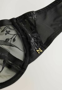 Czarny stanik koronkowy z kwiecistym haftem i siateczkowymi detalami, z złotym akcentem w postaci litery "N" na boku. Miękka tekstura i delikatny design.