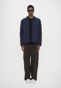 Navy shirt jacket with a pocket, paired with black T-shirt and brown cargo pants. Model wears black sunglasses and white sneakers.