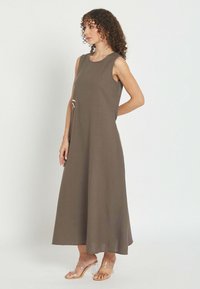 Mujer con cabello rizado que lleva un vestido midi sin mangas de color verde oliva y sandalias de tacón con tiras transparentes, de pie frente a un fondo claro y liso.