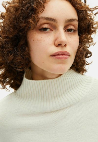 Maglione a collo alto a coste beige chiaro presenta un colletto alto, una morbida texture in maglia ed è mostrato dalla vita in su su un modello con capelli ricci.