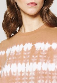 Tričko s krátkým rukávem a kulatým výstřihem v broskvové barvě s batikovým vzorem tvořeným bílými nepravidelnými tvary, vyrobené z měkké bavlněné látky.