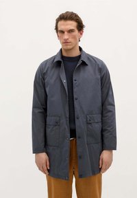 Mørkegrå vandtæt jakke med knaplukning, to frontlommer og krave, båret over en marineblå struktureret sweater og brune bukser.