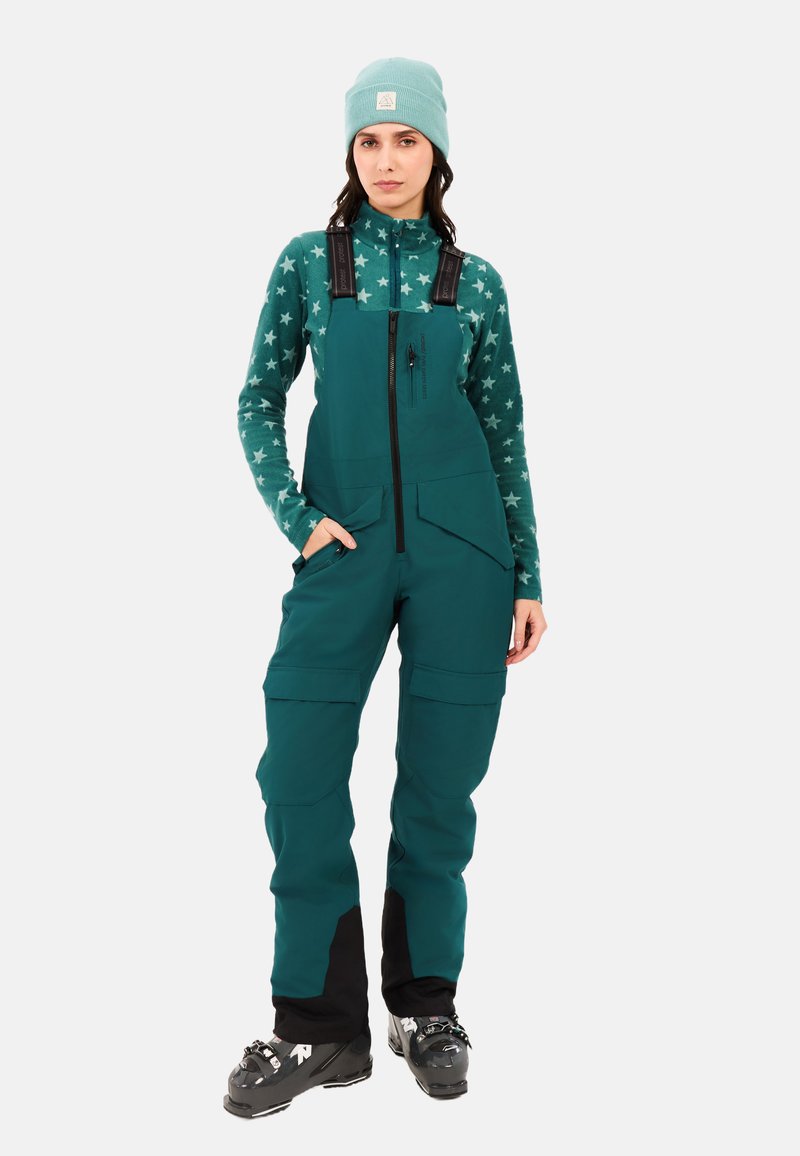 Teal skibroek met verstelbare banden, meerdere zakken en zwarte accenten bij de enkels, gecombineerd met een bijpassende beanie en een fleece met sterpatroon.