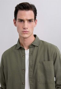 Camisa de botones verde oliva en una tela texturizada con un bolsillo en el pecho y una camiseta blanca de cuello redondo debajo. Diseño de cuello sencillo.