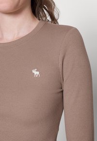 Maglione marrone a maniche lunghe con scollo rotondo e piccolo ricamo di un alce bianco sul petto. Tessuto liscio, design aderente.