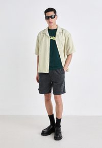 Light yellow, short-sleeve, striped shirt over a dark green t-shirt. Black corduroy shorts and black slip-on shoes complete the outfit.