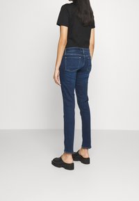 Dark blue slim-fit jeans with a mid-rise waist and back pockets, paired with black shoes. Fabric is denim with a smooth texture.