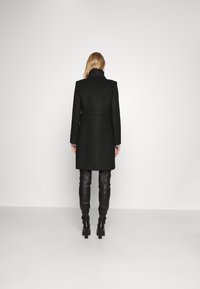 Cappotto di lana nera con collo alto, vita aderente e maniche lunghe, abbinato a pantaloni in pelle nera e tacchi. Texture liscia, design minimalista.