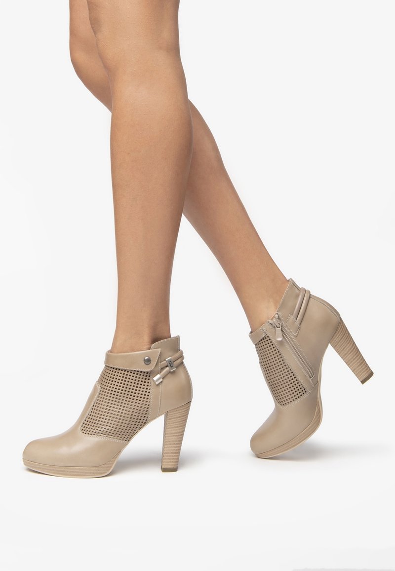 Bottines beige à talons hauts avec panneaux en mesh, fermetures éclair latérales et sangles décoratives portées sur des jambes croisées sur un fond uni.