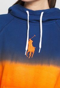 Hedvábná mikina s modro-oranžovým přechodem, bílými šňůrkami a vyšívaným oranžovým logem polo hráče na hrudi.