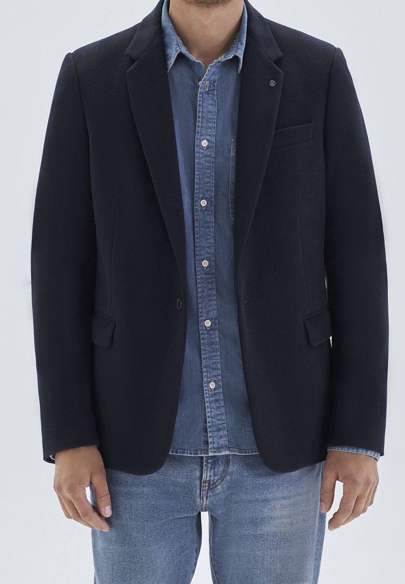 Man wearing dark navy blazer over a buttoned denim shirt and light blue jeans, standing against a plain light background.