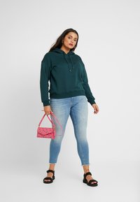 Mørk grønn hettegenser med en frontlomme, kombinert med lyseblå skinnyjeans, svarte sandaler og en liten rosa håndveske.