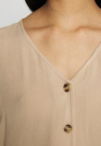 Beige, leichte Bluse mit V-Ausschnitt und drei Knöpfen vorne, mit glatter Struktur und dezentem Glanz.