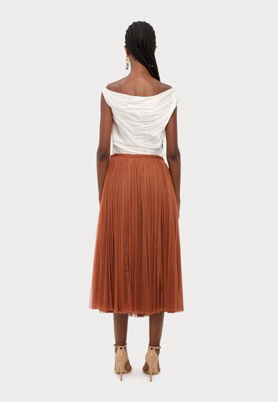 Top bianco off-shoulder con tessuto drappeggiato, gonna plissettata marrone e sandali con tacco beige. La gonna ha un'aspetto testurizzato.