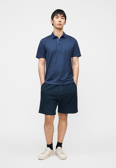 Πουκάμισο πόλο σε ναυτικό μπλε χρώμα με γιακά και τρία κουμπιά, σε συνδυασμό με σορτς σε ίδιο χρώμα. Το μοντέλο φοράει ανοιχτόχρωμα αθλητικά παπούτσια και σκοτεινές κάλτσες.
