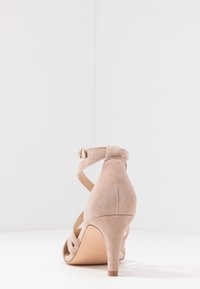 Sandal de tacón alto de ante beige con correa al tobillo y hebilla, exhibida sobre una superficie blanca con un fondo blanco.