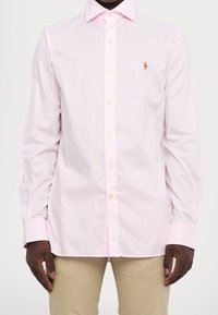 Světle růžová pruhovaná košile s knoflíkovým límcem a dlouhými rukávy. Obsahuje malé logo na hrudi a kulaté manžety.
