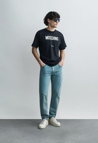 Must Moschini särk valge logoga, kombineeritud helesiniste sirgelõikeliste teksade ja valgete tossudega. Modell seisab neutraalses keskkonnas.