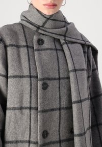 Casaco de lã cinza com um padrão de xadrez preto. Apresenta um colarinho alto, fechos de botão e uma textura leve. Design semelhante a um cachecol na gola.