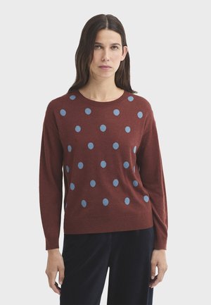 Mujer con cabello largo y oscuro, vistiendo un suéter granate con lunares azules y pantalones negros sobre un fondo blanco liso.