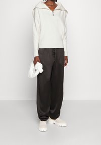 Witte sweater met rits en capuchon, gecombineerd met zwarte wijde broekspijpen en chunky witte enkellaarsjes. Overal gladde, zachte texturen.