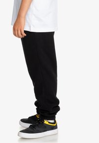 Svarta fleece-joggingbyxor med elastiska muddar, parat med svarta sneakers med gula accenter och vit sula. En vit t-shirt är synlig.