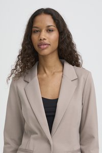 Mujer con cabello largo y rizado que lleva una chaqueta beige sobre una blusa negra, de pie frente a un fondo claro y simple.