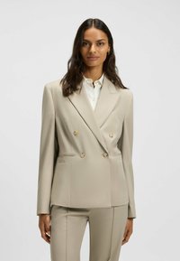 Beige double-breasted blazer with two front pockets, gold-tone buttons, and a notched collar, made from smooth, structured fabric.