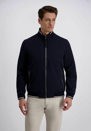 Man draagt een marineblauwe jas met rits en witte bies op de kraag en manchetten, gecombineerd met lichtbeige broek, staand tegen een effen achtergrond.