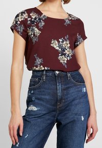 Krátký rukáv vínově červená květinová blůza s krémovými, modrými a šedými květinami, kombinovaná s roztrhanými vysokými džíny s tmavým umytím.