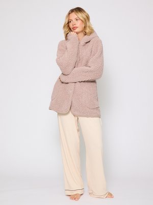 Giacca oversized di colore rosa chiaro, morbida e con cappuccio, dotata di tasche frontali, abbinata a pantaloni beige larghi con rifiniture nere.