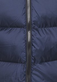 Marinblå puffjacka med horisontell quiltning. Utrustad med en svart dragkedja med ett silverfärgat handtag. Materialet verkar slätt och lätt.