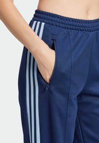 Ruka v náručí modrých športových nohavíc so svetlomodrými bočnými pruhmi a vreckom na zips, zobrazujúce elastický pás a spodnú časť trupu.