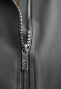Dark gray zip-up jacket close-up, featuring a smooth fabric texture and silver metal zipper with "self" branding on the pull tab.
