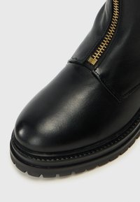 Stivaletto nero in pelle con una texture liscia, punta rotonda e dettagli in zip dorata laterale. Suola in gomma spessa con un motivo di battistrada.
