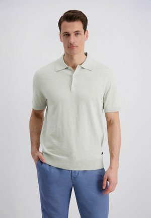 Mann trägt ein hellgraues kurzärmeliges Polohemd und blaue Hose und steht vor einem einfarbigen weißen Hintergrund.
