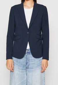 Mörkblå kavaj i slätt tyg, med en knapp, uppslagna slag och två framfickor. Kombinerad med en vit topp och ljusa jeans.