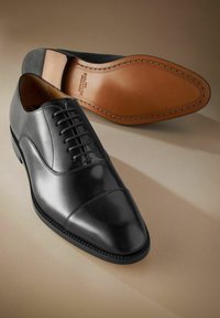 Zapato oxford de cuero negro con puntera puntiaguda, cordones en la parte delantera y suela de cuero marrón claro en contraste. Detalles de costura sutil a lo largo de los bordes.