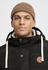 Doudoune noire avec une doublure douce couleur crème, présentant un patch logo marron et des cordons de serrage blancs au niveau de la capuche. Bonnet en tricot marron.