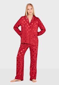 Conjunto de pijama de satén rojo con un gran patrón de lunares. Presenta una camisa con botones y pantalones de corte holgado, textura suave, mangas largas y un bolsillo en el pecho.
